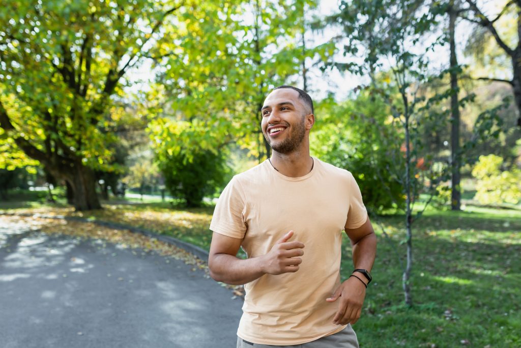 Cheerful and successful hispanic man jogging in the park, man running on a sunny day, smiling and happy having an outdoor activity.